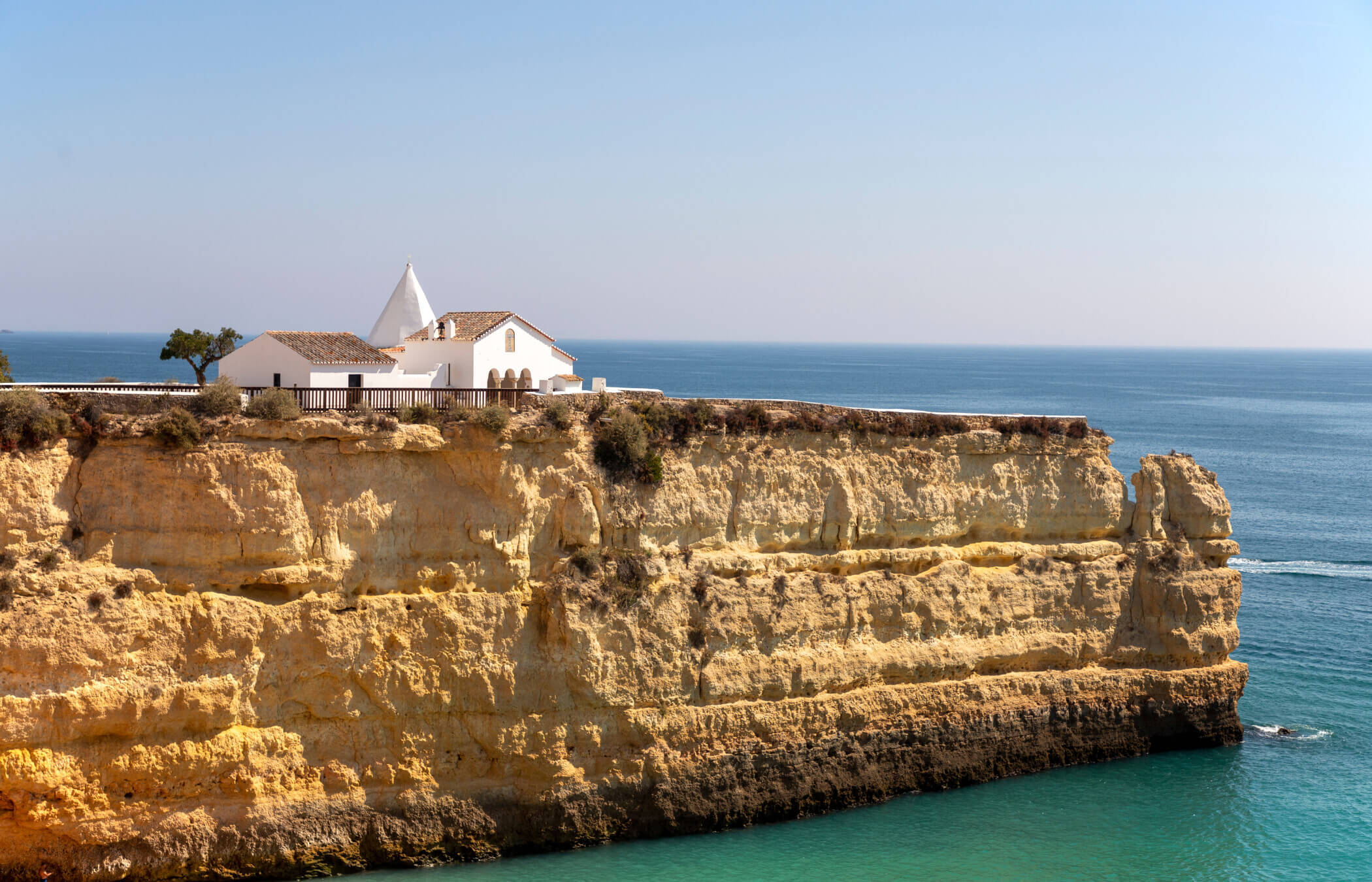 A white church sits on top of a golden cliff, surrounded by blue ocean. This is Nossa Senhora da Rocha (Our Lady of the Rock) in the Algarve, Portugal.