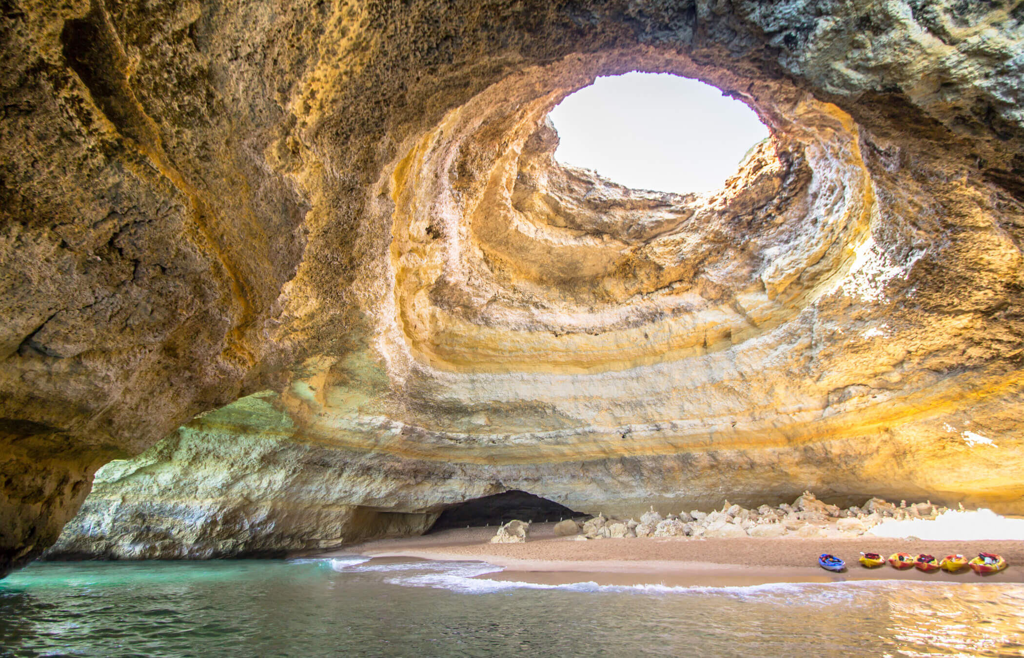 Aome kayaks sit on a small beach with water creeping up on the shore inside a large cave with a large 'skylight' illuminating inside of the cave. This is Benagil Cave in Portugal.