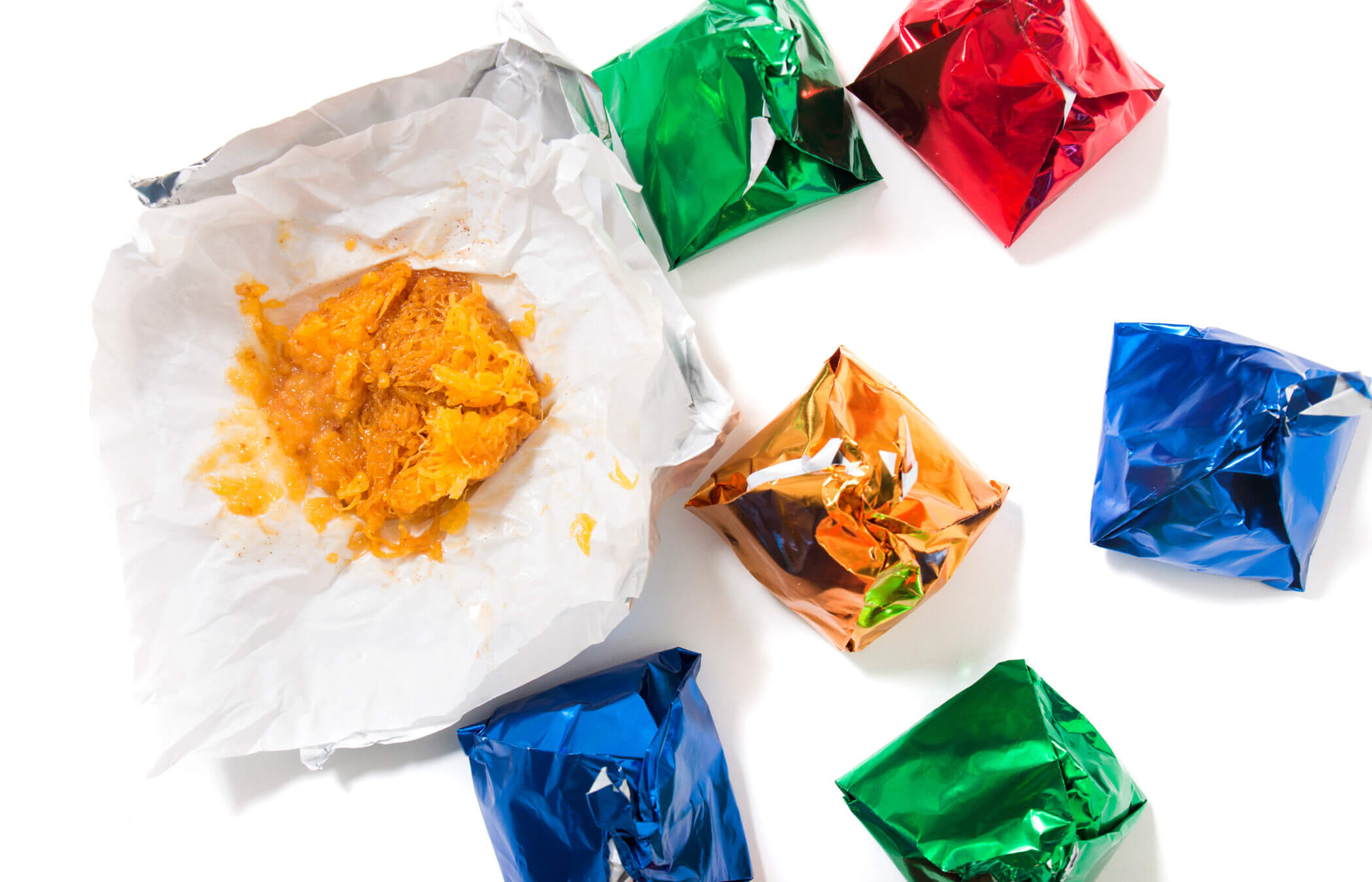 Seven 'Dom Rodrigo' desserts sit on a white background. Each is wrapped in a colourful foil - green, blue, red and orange - bar one, which is open, revealing a mushy orange substance.
