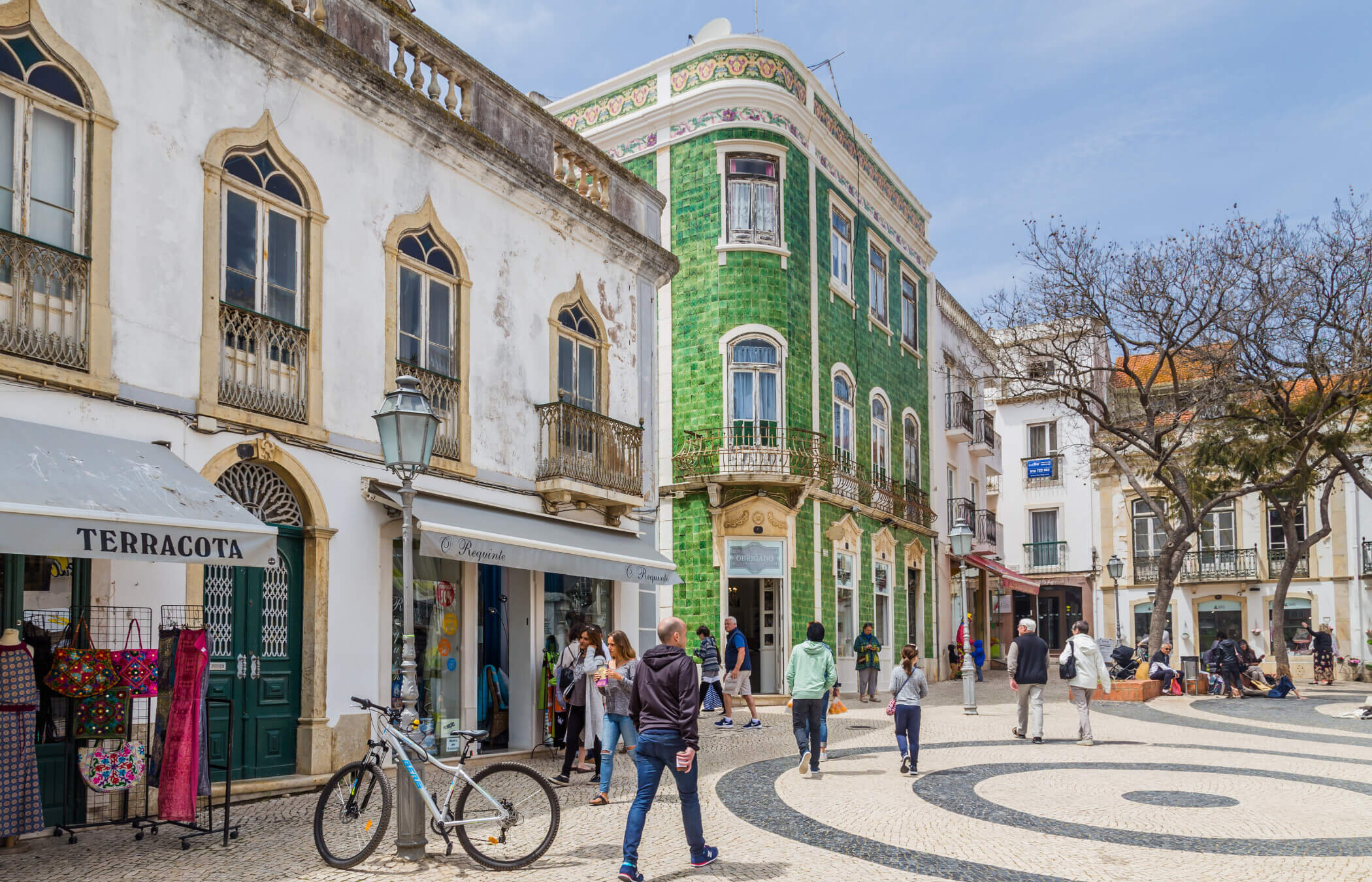 A vibrant town square with swirling patterned tiles on the ground, featuring a striking green building adorned with intricate ornamental design. Jacaranda trees tower over park benches while people sit and walk around, enjoying the atmosphere.