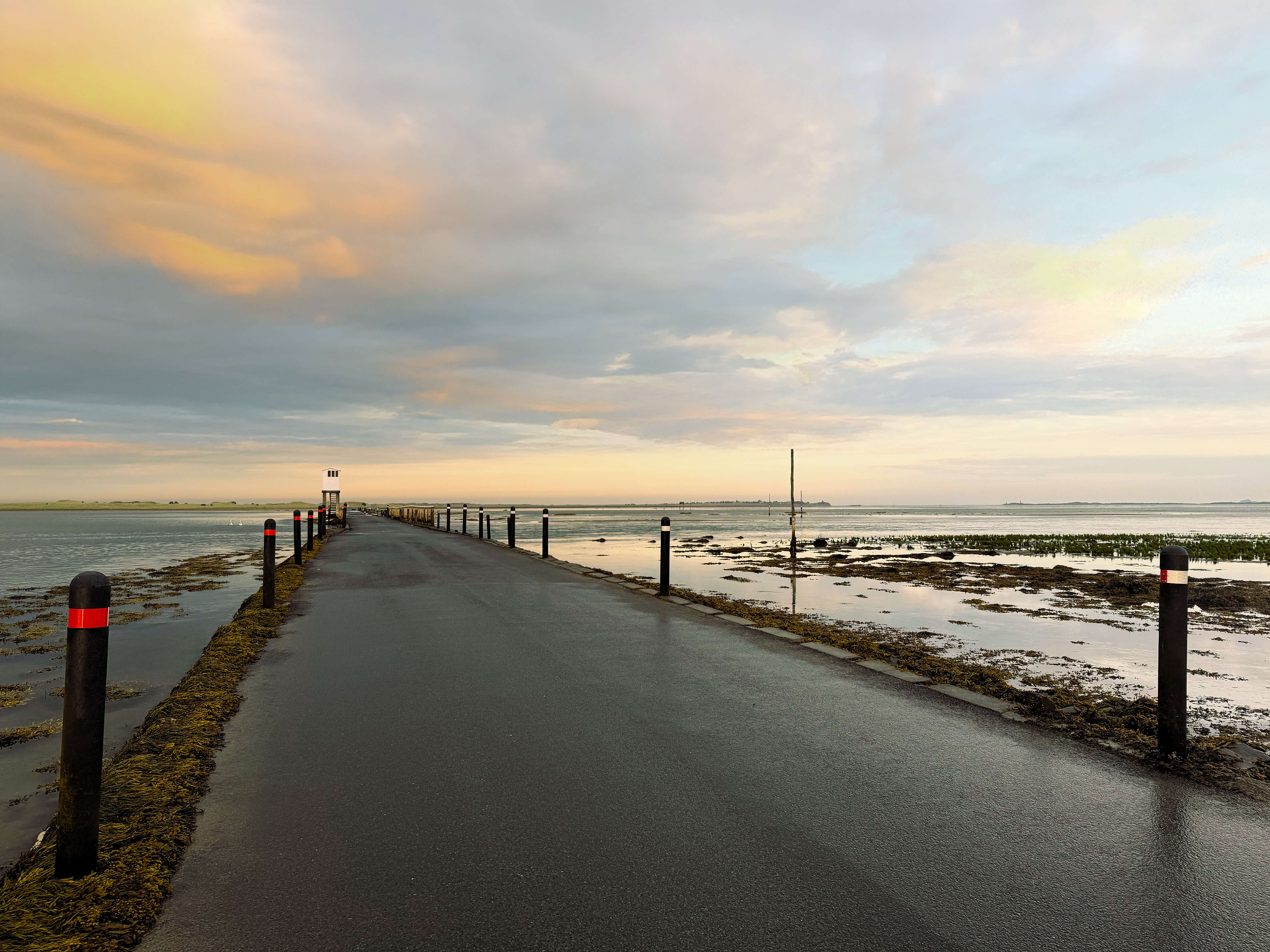 The road crossing over to Lindisfarne. We see the pilgrim path to the side, with the markers that show the depth of the tide. Holy Island sits in the haze of the background. The sky is pleasant but cloudy, the sun is rising.