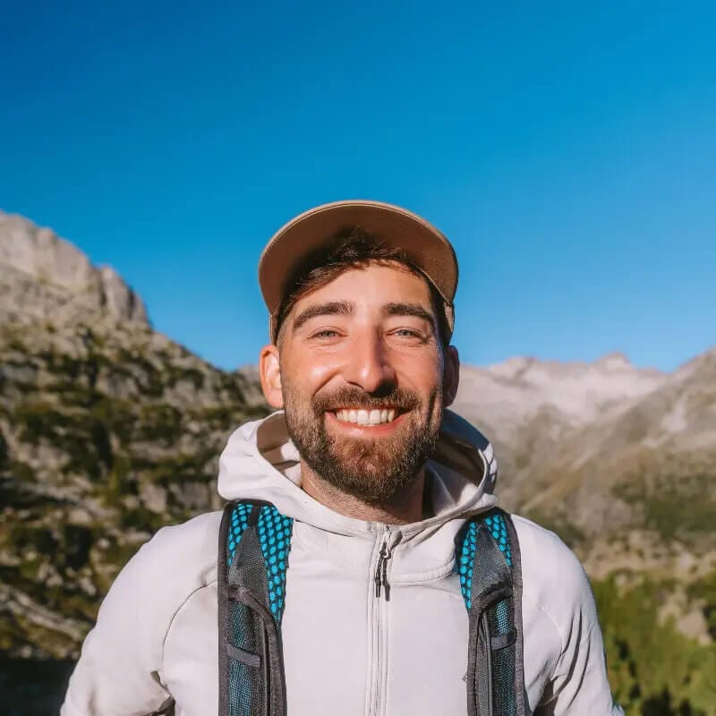 A smiling man wearing a backpack and a grey hoodie looks directly at the camera. He is wearing a beige baseball cap. In the background we can see a rugged outdoor scene.