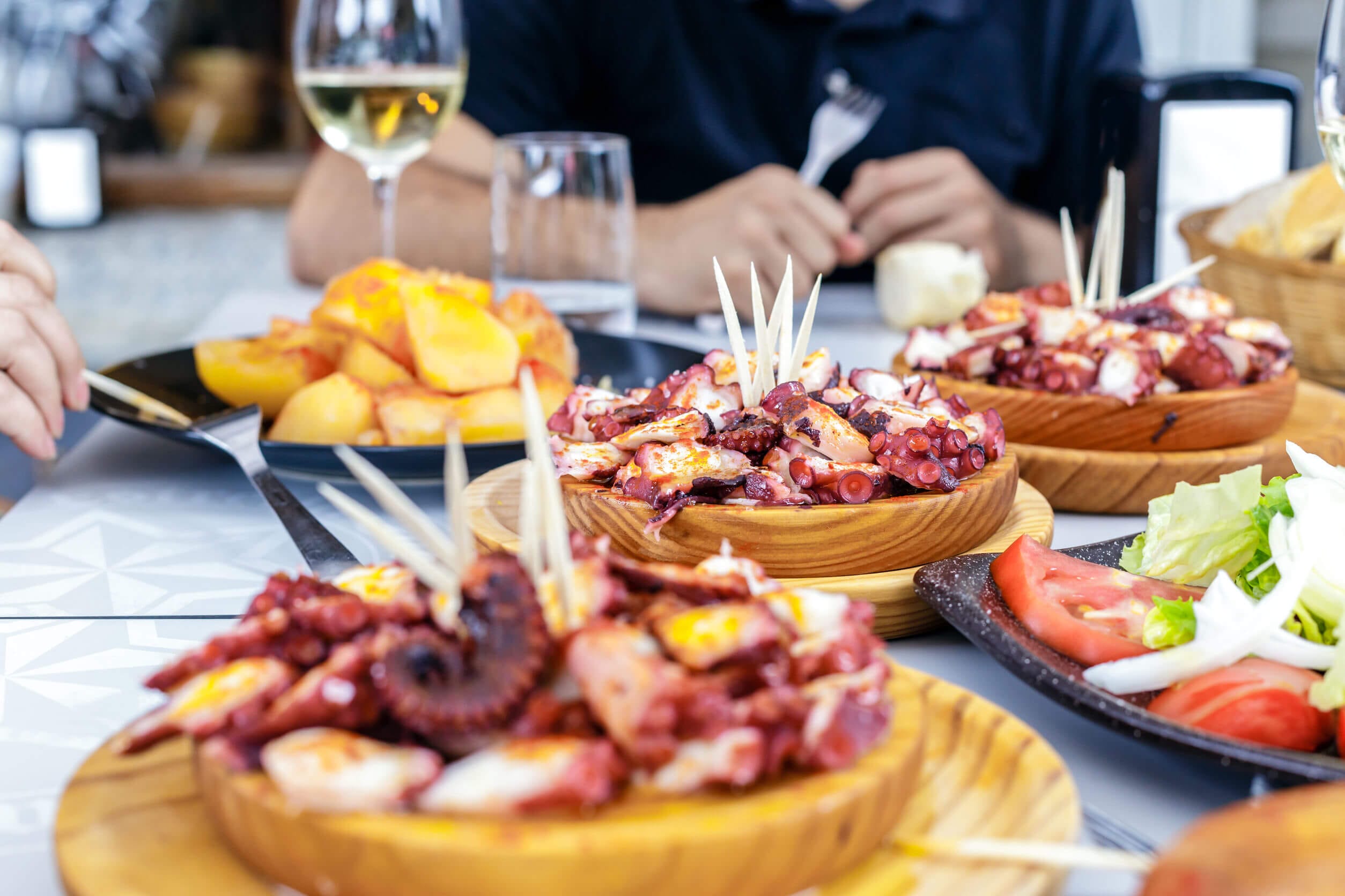A table with 3 plates of pulpo a la gallega (Galician octopus), one with crispy golden brown potatoes and one with a garden salad sit on a table with a white table cloth, along with a glass of white wine and a glass of water. In the background a man in a navy teeshirt holds a fork in his right arm