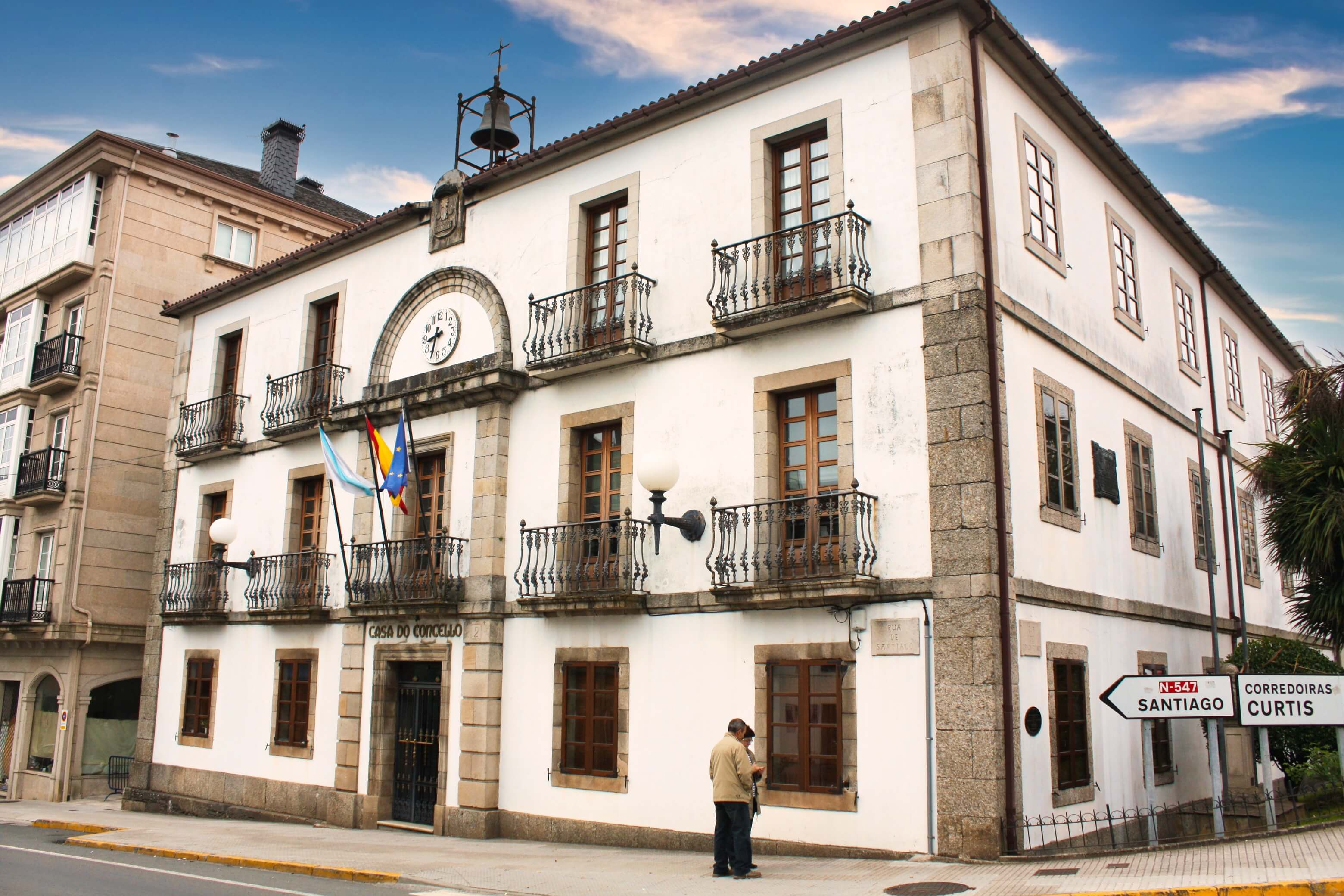Casa do Concello (town hall) in Arzúa where Camino Primitivo merges with Camino Francés. Three-storey white building with grey granite corners and window surrounds. Wrought iron balconies on upper floors. Bell tower with clock rises from roofline topped with iron cross. Galician and Spanish flags hang from balconies. Elderly pilgrim with walking stick stands at entrance. Road sign indicates "Santiago" direction.
