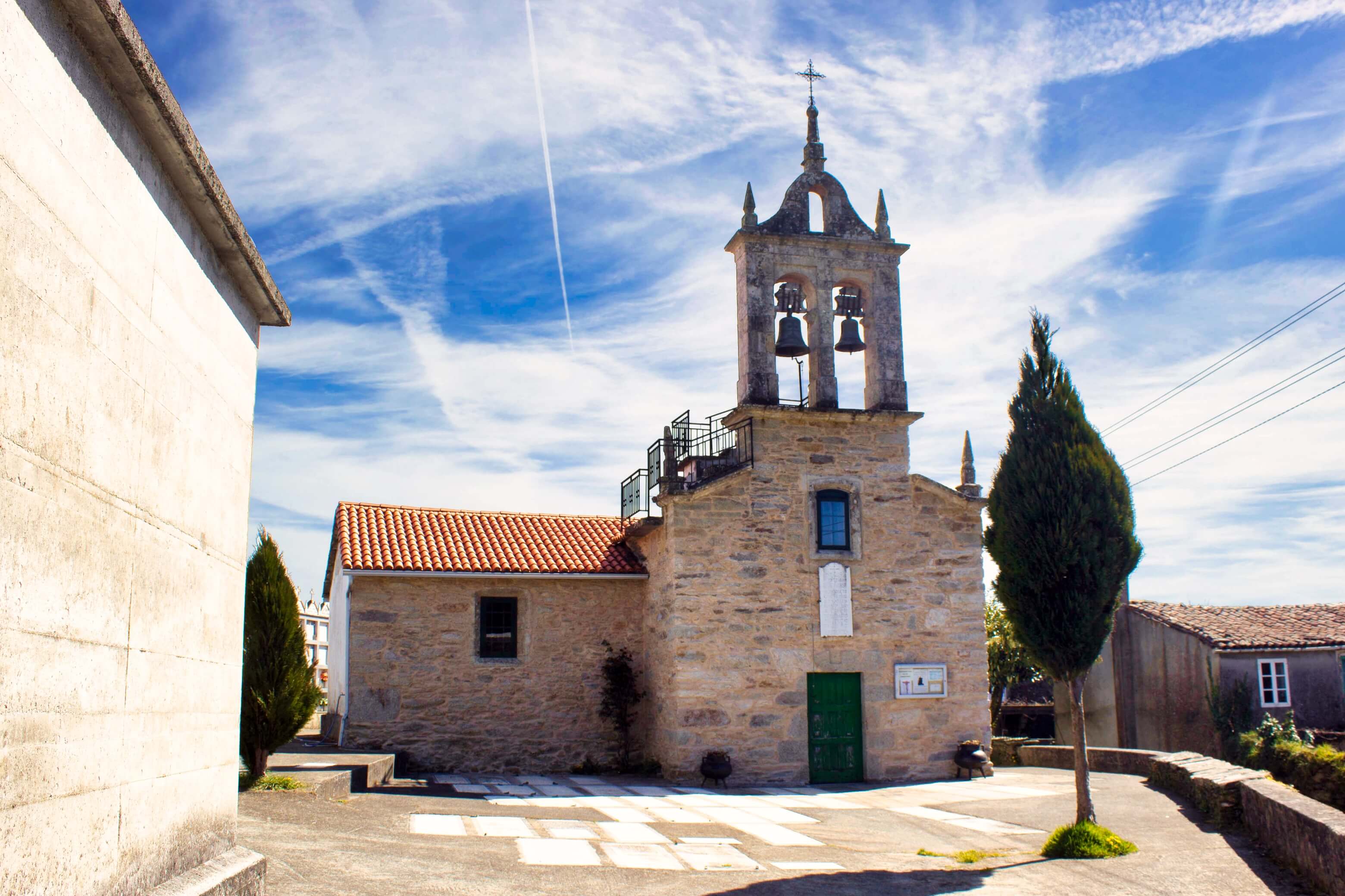 Small stone village church with a bell tower holding two bells and a cross on top. The church has a green door, red tile roof, and stands in a paved square with a slender tree under a bright blue sky with wispy clouds in Arzúa.