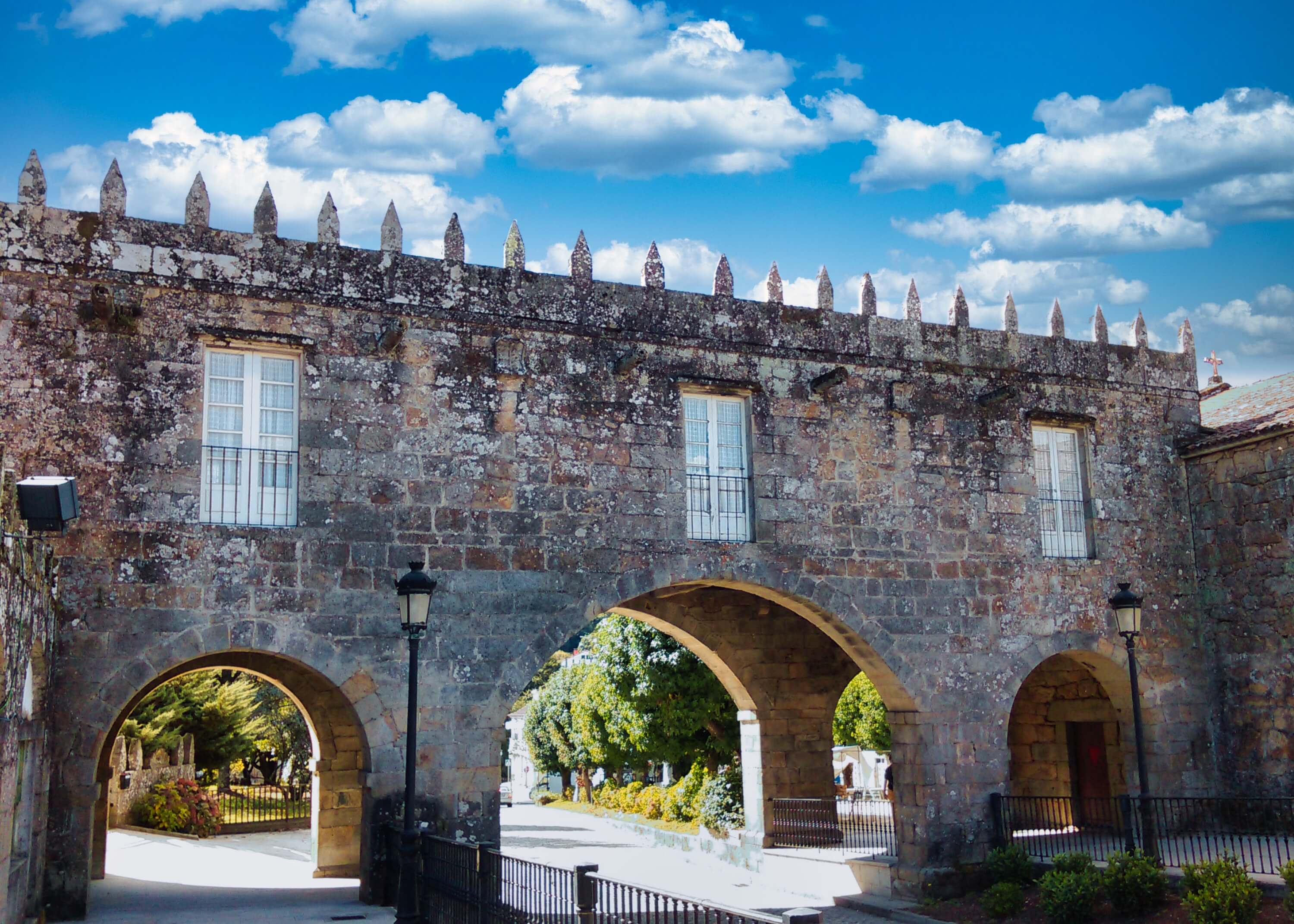 A medieval stone building with three arched gateways and battlemented parapet, with traditional lamp posts and green trees visible through the arches, Negreira, Galicia.