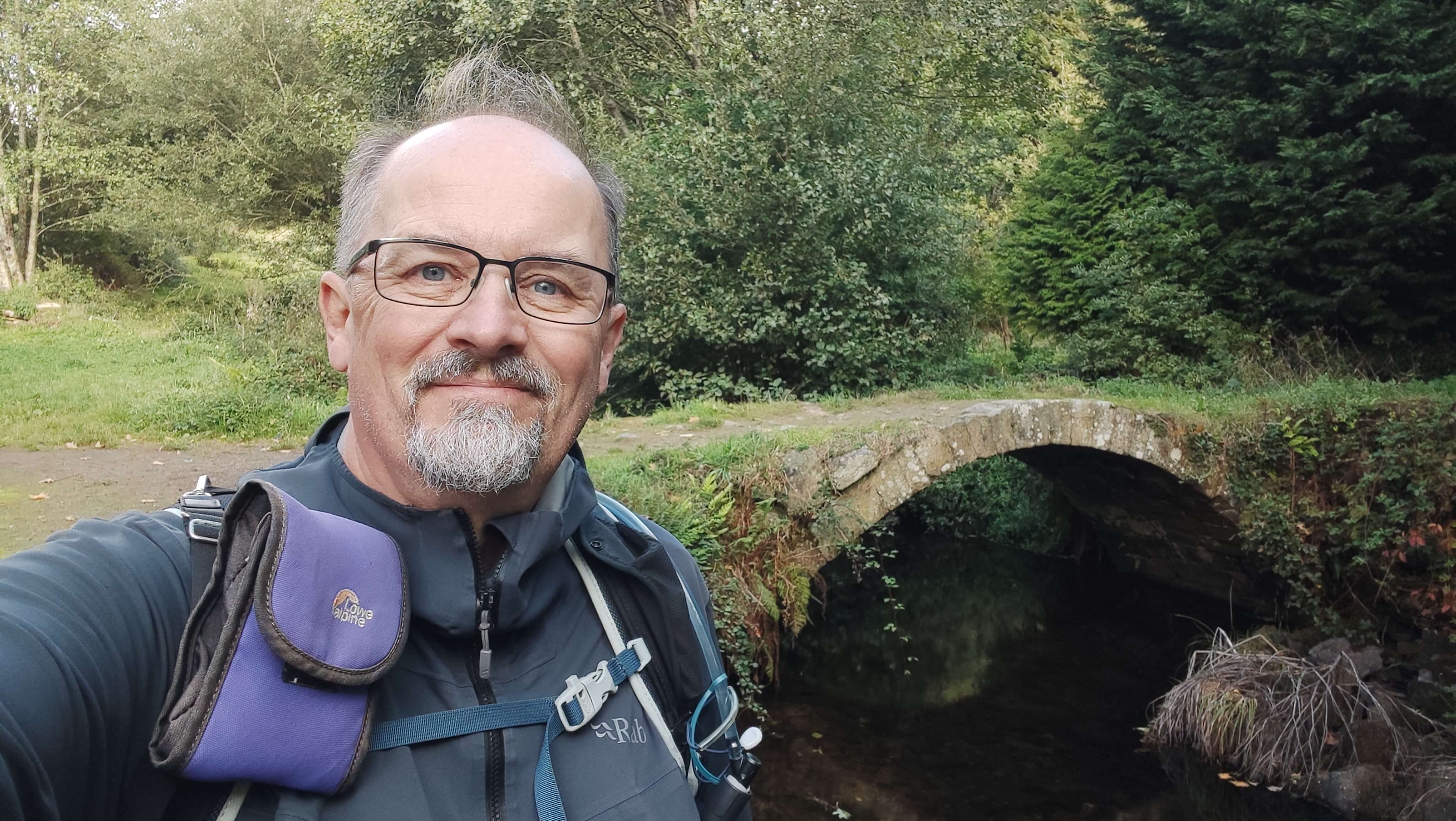 Close-up selfie of a smiling male pilgrim with glasses and grey goatee beard standing on the Camino Primitivo in rural Galicia. He wears a grey fleece jacket with backpack straps visible on his shoulders. His warm, contented expression captures the spirit of the pilgrimage. Behind him stretches a beautiful medieval stone bridge with a single rounded arch spanning a narrow stream, its weathered grey stones covered in patches of moss and trailing vegetation. The dark water below reflects the arch creating a perfect circle. Lush green grass slopes lead down to the riverbank on both sides, while dense mixed woodland of deciduous trees and evergreens creates a natural canopy in the background. The peaceful rural scene, typical of the Galician countryside along the Camino Primitivo, shows the kind of ancient infrastructure that has served pilgrims for centuries on their journey to Santiago de Compostela."