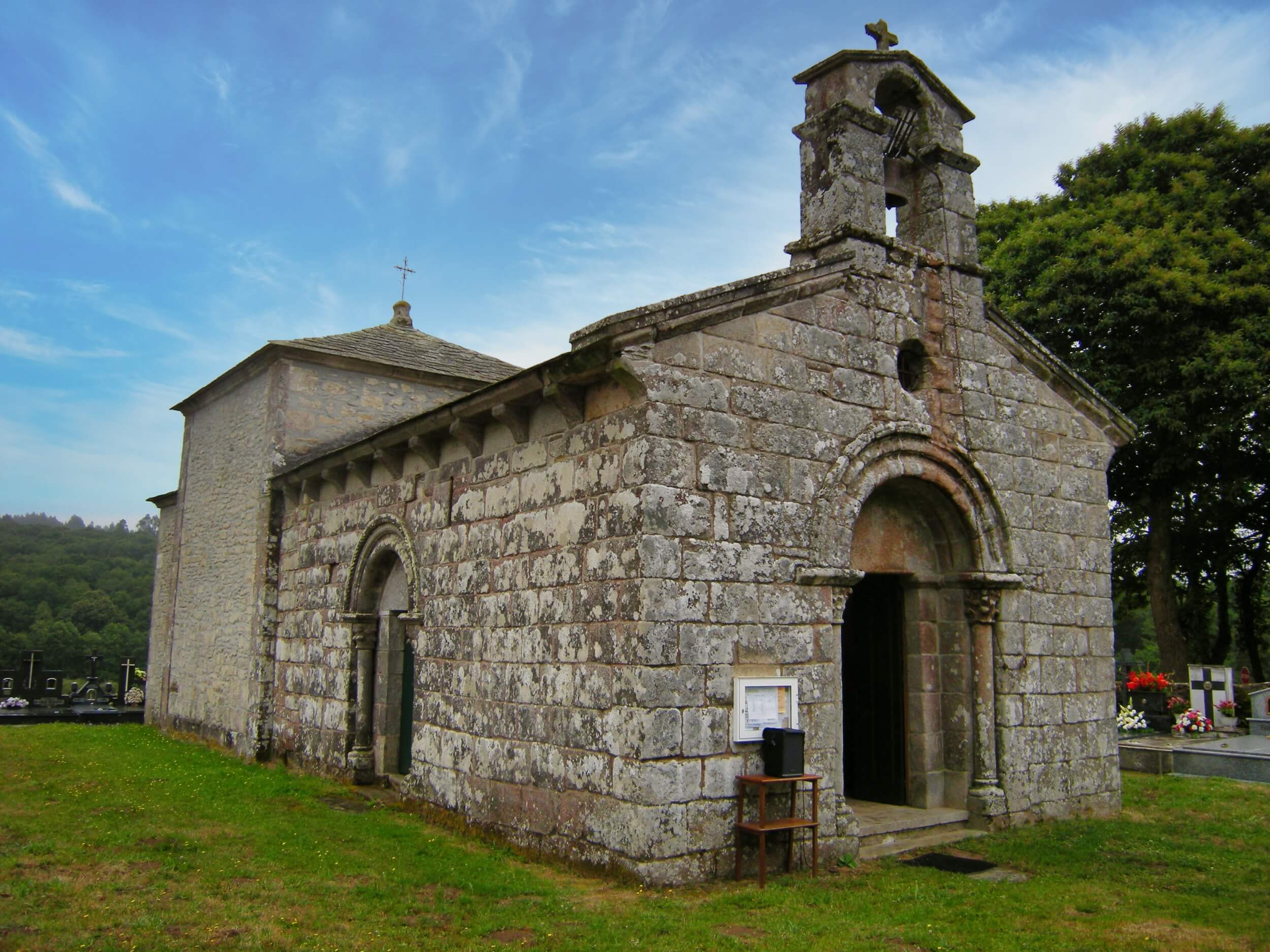 Small rural Romanesque stone church with arched doorways and a simple bell gable, set in a grassy cemetery with headstones. The church stands under a blue sky with trees and hills in the background.